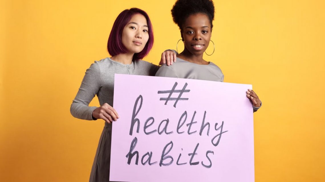 Two women holding a sign with '#healthy habits' on a vibrant yellow background.