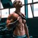 Muscular man showing abs and tattoos in a gym setting.